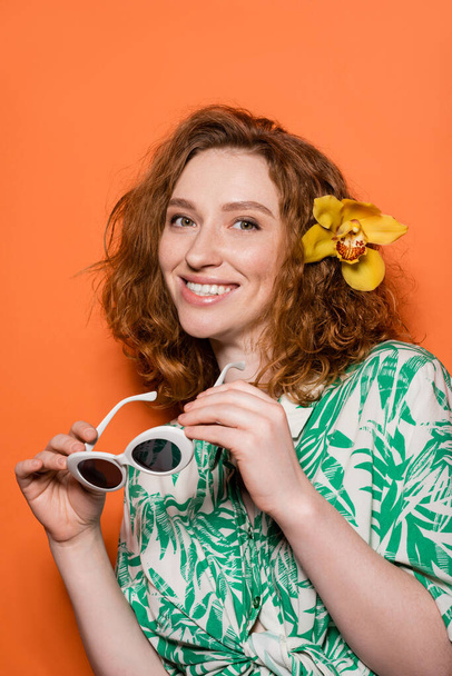 Mujer de pelo rojo joven positivo con flor de orquídea en el pelo mirando a la cámara y sosteniendo gafas de sol y de pie sobre fondo naranja, verano casual y concepto de moda, Cultura Juvenil - Foto, Imagen