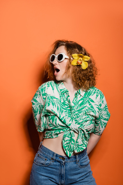Shocked redhead woman with sunglasses and orchid flower wearing blouse with floral print and jeans while posing and standing on orange background, summer casual and fashion concept, Youth Culture - Photo, Image