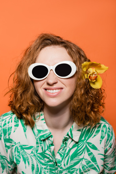 Usmívající se mladá žena s orchidej květiny v rudých vlasech nosí sluneční brýle a blůzu s květinovým potiskem, zatímco stojí izolované na oranžové, stylové ležérní oblečení a letní vibrace koncept, Mládež kultury - Fotografie, Obrázek