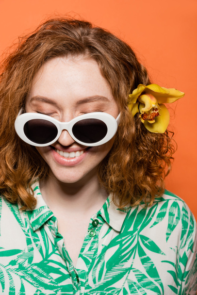 Mujer pelirroja alegre con gafas de sol y flor de orquídea con blusa con estampado floral y de pie aislado en naranja, elegante atuendo casual y concepto de vibraciones de verano, Cultura Juvenil - Foto, Imagen