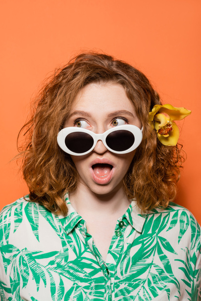 Jeune femme choquée avec fleur d'orchidée en cheveux roux portant des lunettes de soleil et regardant loin tout en posant et debout sur fond orange, concept décontracté et mode d'été, Culture de la jeunesse - Photo, image