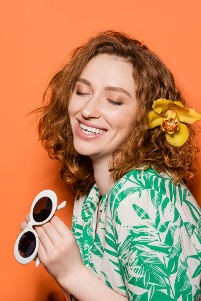 Positives junges Rotschopf-Model mit Orchideenblüte im Haar mit Sonnenbrille und Bluse mit modernem Blumenmuster auf orangefarbenem Hintergrund, Sommer-Freizeit- und Modekonzept, Jugendkultur - Foto, Bild