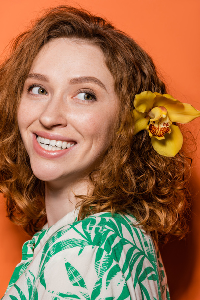 Πορτρέτο της χαρούμενης νεαρής και κοκκινομάλλας γυναίκας με ορχιδέα λουλούδι στα μαλλιά κοιτάζοντας μακριά και ποζάροντας σε μπλούζα, ενώ στέκεται σε πορτοκαλί φόντο, καλοκαίρι casual και μόδα έννοια, Νεολαία Πολιτισμού - Φωτογραφία, εικόνα