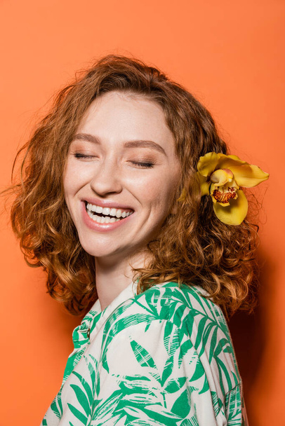 Счастливая молодая женщина с цветком орхидеи в рыжих волосах и модная блузка с цветочным принтом стоя с закрытыми глазами на оранжевом фоне, летняя повседневная и модная концепция, молодежная культура - Фото, изображение
