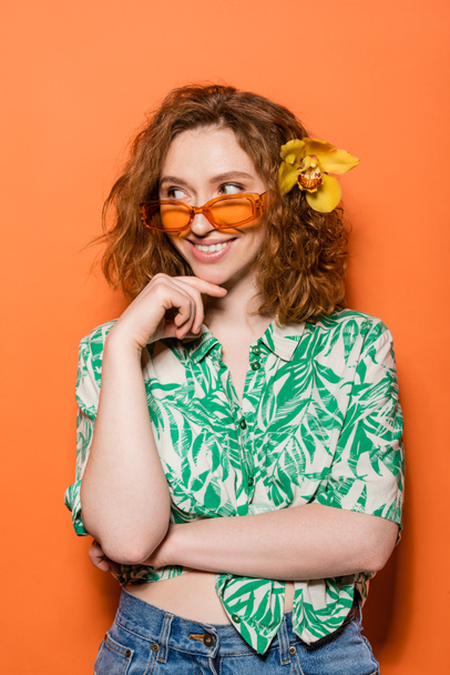 Cheerful young woman with orchid flower in hair wearing sunglasses and blouse with floral pattern while standing on orange background, summer casual and fashion concept, Youth Culture - Photo, Image