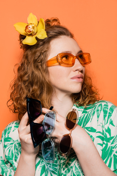 Втомлена молода жінка з квіткою орхідеї в волоссі тримаючи сонцезахисні окуляри і озираючись, виставляючи і стоячи на помаранчевому тлі, літні випадкові і модні концепції, Молодіжна культура - Фото, зображення