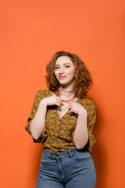Fashionable and redhead woman in blouse with abstract pattern and jeans touching necklaces and smiling on orange background, stylish casual outfit and summer vibes concept, Youth Culture - Φωτογραφία, εικόνα