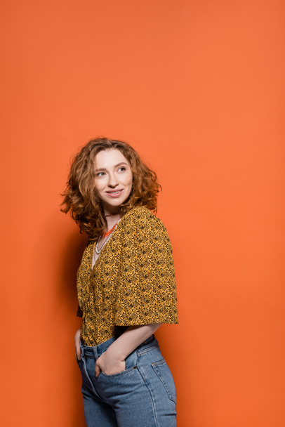 Joyful young redhead woman in blouse with abstract pattern holding hands in pockets of jeans and looking away on orange background, stylish casual outfit and summer vibes concept, Youth Culture - Фото, изображение