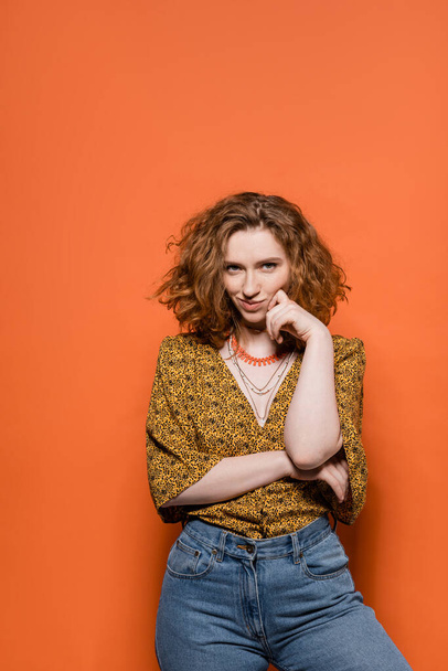 Confident and smiling red haired woman in blouse with abstract pattern and jeans looking at camera while standing on orange background, stylish casual outfit and summer vibes concept, Youth Culture - Photo, Image