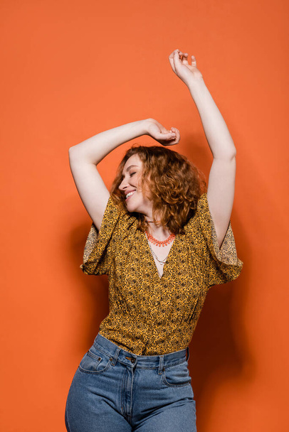 Cheerful young red haired woman in yellow blouse with abstract pattern and jeans dancing while standing on orange background, stylish casual outfit and summer vibes concept, Youth Culture - Photo, Image