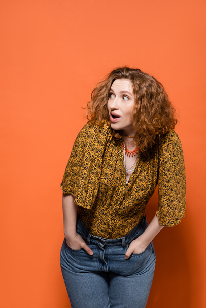 Shocked young red haired woman in yellow blouse and trendy jeans looking away while posing and standing on orange background, stylish casual outfit and summer vibes concept, Youth Culture - Photo, Image