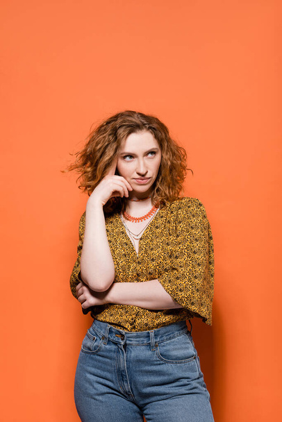 Pensive young red haired woman in modern yellow blouse and jeans looking away while standing and posing on orange background, stylish casual outfit and summer vibes concept, Youth Culture - 写真・画像