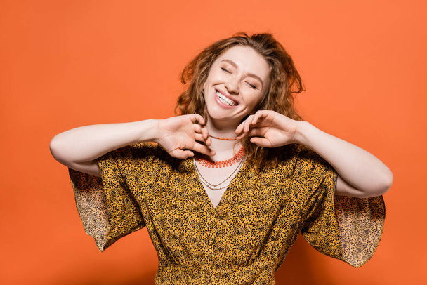 Positive young red haired model in yellow blouse with abstract pattern touching necklaces and standing with closed eyes on orange background, stylish casual outfit and summer vibes concept, Youth Culture - 写真・画像