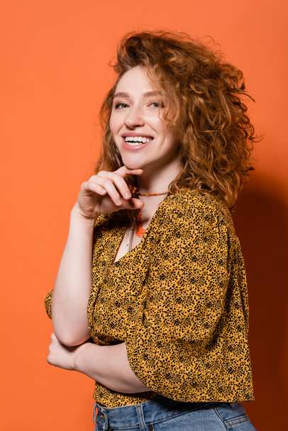 ネックレスの幸せな若い赤い髪の女性、カメラを見て、オレンジの背景にポーズを抽象的なパターンを持つ黄色のブラウス、スタイリッシュなカジュアルな衣装と夏のバイブのコンセプト、若者文化 - 写真・画像