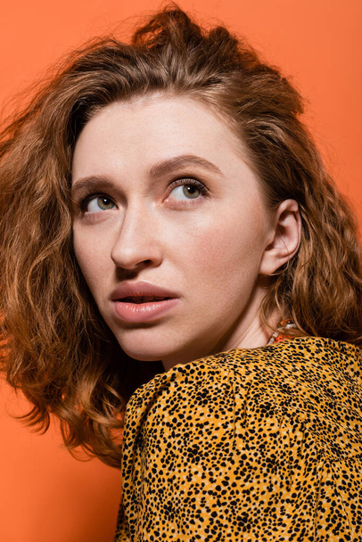 Portrait of young red haired woman with no-makeup look wearing yellow blouse with abstract pattern and looking away isolated on orange, stylish casual outfit and summer vibes concept, Youth Culture - Φωτογραφία, εικόνα