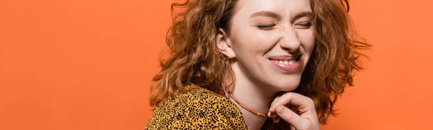 Pleased young red haired woman with natural makeup touching necklace and posing in blouse with abstract pattern isolated on orange, stylish casual outfit and summer vibes concept, banner - Valokuva, kuva