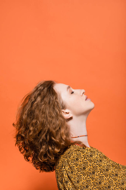 Side view elegáns fiatal vörös hajú nő természetes smink blúz modern absztrakt nyomtatott álló elszigetelt narancs, elegáns alkalmi ruhát és nyári vibrálás koncepció, Ifjúsági Kultúra - Fotó, kép