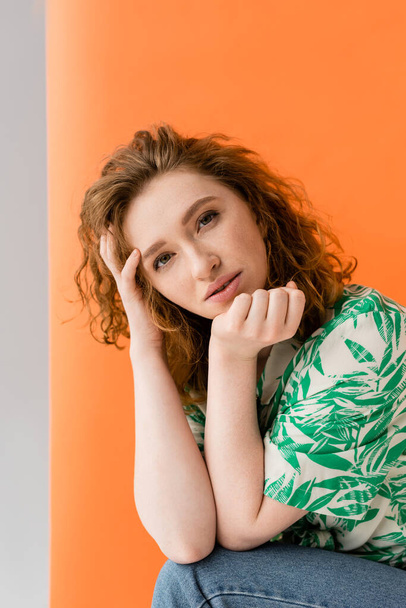 Portrait of young redhead woman in blouse with floral pattern and jeans looking at camera while standing on orange and grey background, trendy casual summer outfit concept, Youth Culture - Photo, image