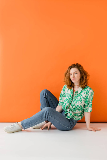 Full length of young red haired woman with natural makeup posing in blouse with floral pattern and jeans while sitting on orange background, trendy casual summer outfit concept, Youth Culture - Photo, Image