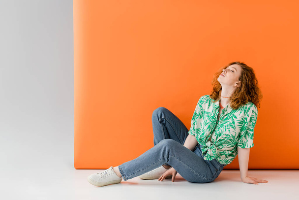 Mujer de pelo rojo joven y relajada con los ojos cerrados en blusa con estampado floral y jeans sentados sobre fondo gris y naranja, concepto de atuendo de verano casual de moda, cultura juvenil - Foto, Imagen