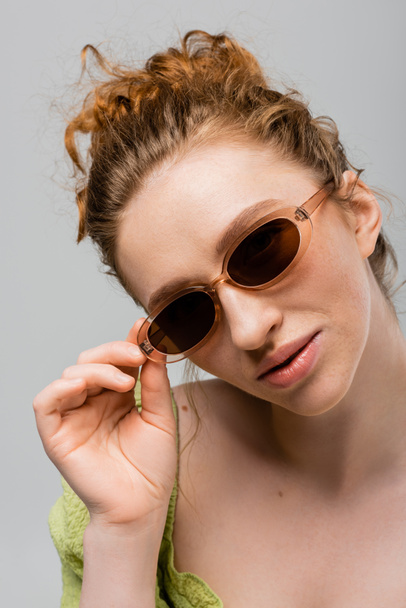 Portrait of confident, red haired and freckled woman in green blouse touching sunglasses while posing and standing isolated on grey, trendy sun protection concept, Youth Culture - Photo, Image