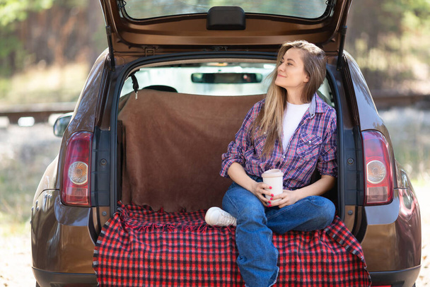 街の騒音から離れて休息している。自然の中で精神的な健康と孤独。一人で休む-若い女性は自然の中で彼女の車のトランクの中でコーヒー休憩を飲みます. - 写真・画像