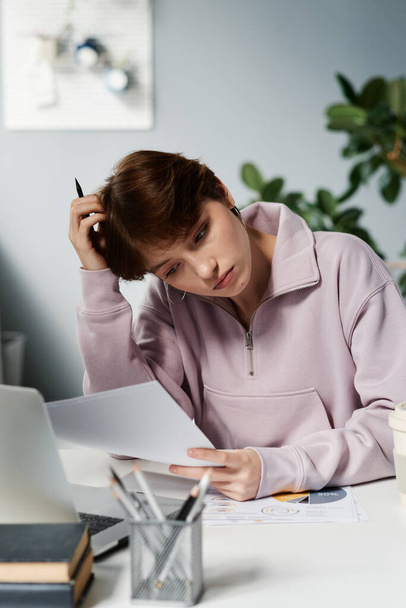 Puzzled young female manager in sweatshirt sitting at desk and reading papers while analyzing data in office - Φωτογραφία, εικόνα