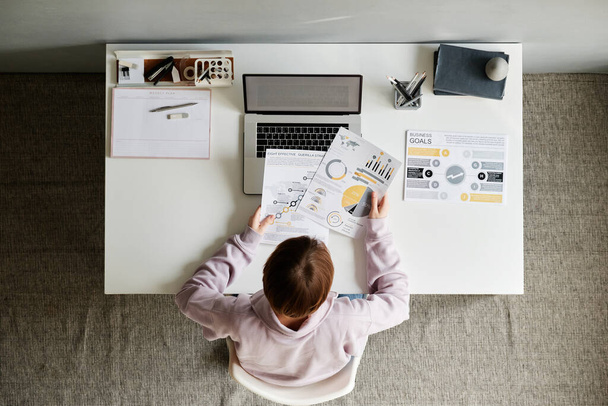 High angle view of short-haired woman in sweatshirt sitting at desk and working with papers while thinking about business strategy - Фото, изображение