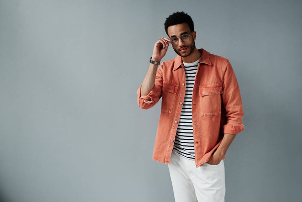 Portrait of content stylish young African American man in coral shirt holding hand in jeans pocket and adjusting glasses - Foto, Imagen