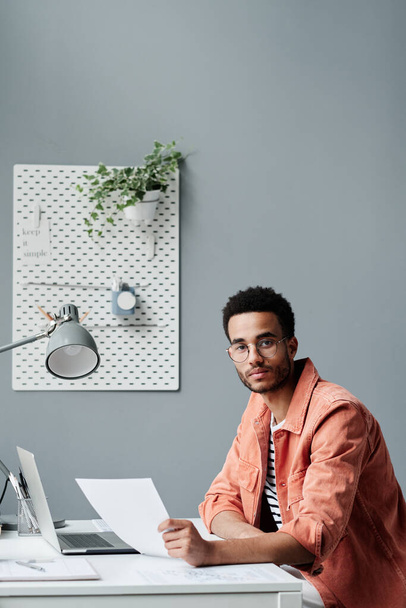 Portrait of serious young black business specialist in casual shirt sitting in office with gray wall and analyzing paper - Fotoğraf, Görsel