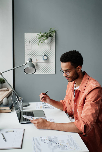 Serious young Black businessman in bright shirt sitting at desk and examining papers in office - 写真・画像