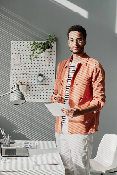 Portrait of content handsome young black manager standing with paper at desk in modern office - 写真・画像