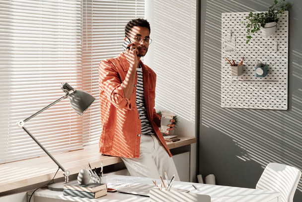 Portrait of confident handsome young black man in stylish outfit standing with takeout coffee at window and chatting by phone in office - Valokuva, kuva