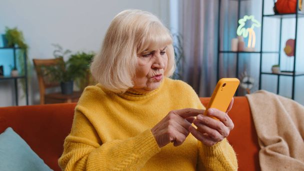 Smutne niezadowolona babcia używać smartfona wpisując przeglądanie, traci coraz zaskakujące nagłe wyniki loterii, złe wieści, utrata fortuny, niepowodzenie, termin, wirus, dług. Starsza kobieta w domu na kanapie - Zdjęcie, obraz