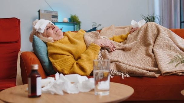 Abuela enferma que sufre de frío o alergia dolor acostado en el sofá en la sala de estar en casa. Mujer anciana enferma con toalla en la cabeza estornuda toallitas mocos en la servilleta. pandemia de cuarentena por Coronavirus - Foto, imagen