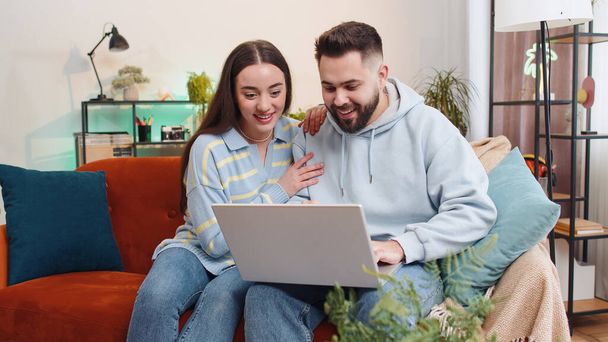 Overjoyed family marriage couple man woman use laptop celebrate great big win good news lottery jackpot, lucky victory, playing game, shopping online at home. Husband and wife together on sofa in room - Foto, imagen