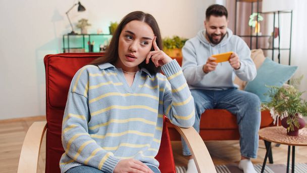 Unhappy woman looking at camera, quarreling with man player addicted of game playing on smartphone at home. Divorce, conflict problem, crisis in married family couple, difficulties. Husband and wife - 写真・画像