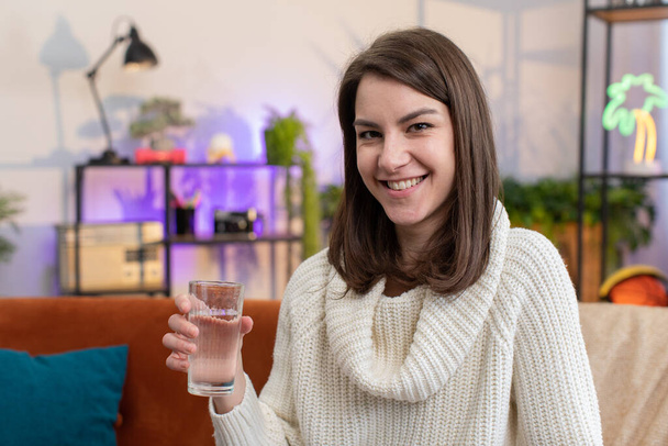 Жаждущая молодая женщина, держащая в руках стакан натуральной воды, делает глотки питьевой воды, предотвращающей обезвоживание, сидящей дома. Девушка с хорошими жизненными привычками, здоровое похудение, концепция потери веса - Фото, изображение
