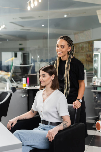 šťastný kadeřník a žena klienta v salonu krásy, veselý kosmetický pracovník s copánky stojící v blízkosti tetované ženy, diskutovat o ošetření vlasů, prodloužení vlasů, spokojenost zákazníka  - Fotografie, Obrázek