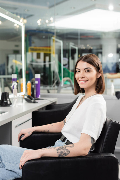 happy female client, tattooed young woman sitting in hairdressing chair and looking at camera in hair salon, beauty service, hair extension, hair coloring, blurred background, generation z  - Photo, Image