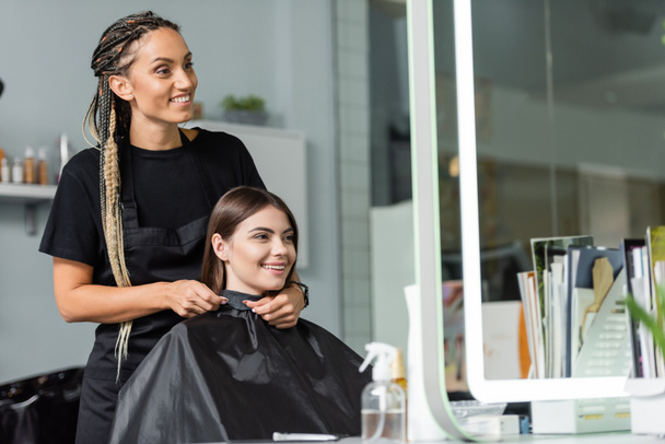 przemysł kosmetyczny, stylista włosów z warkoczami mocowanie peleryna fryzjerska na kliencie żeńskim, przedłużanie włosów, leczenie włosów, klient salon, zawód kosmetyczny, salon pracy, uśmiechając się razem  - Zdjęcie, obraz