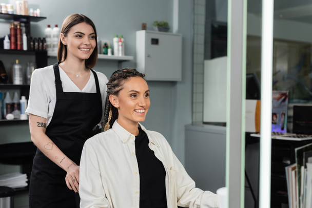 hair professional, happy and tattooed beauty worker in apron and female client with braids smiling in salon, beauty industry, salon job, customer in salon, hairdresser, salon services  - Photo, Image