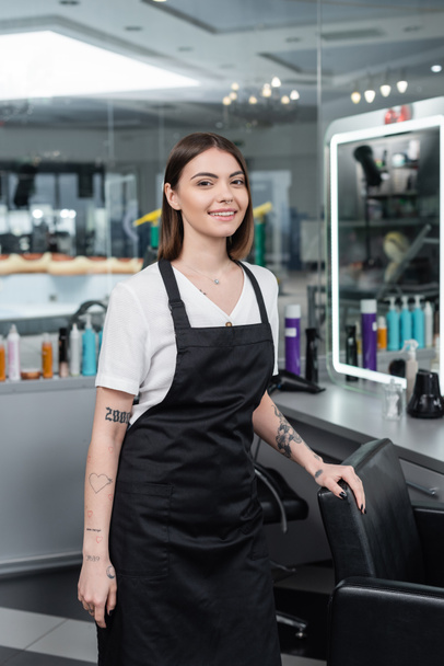 professional headshots, tattooed beauty worker in apron looking at camera in beauty salon, hairdressing chair, hair stylist, hair coloring, salon job, beauty salon work, hair trends  - Photo, Image