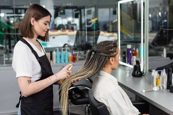 hair salon, tattooed beauty worker in apron applying hair product on braids of woman in salon, spray bottle, happy beauty worker, client satisfaction, salon customer, smile, side view - Photo, Image