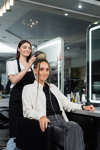 hair make over, cheerful hair stylist doing hair bun to female client with braids, cheerful women, client satisfaction, customer in salon, beauty service, salon experience, feminine, tattooed  - Photo, Image