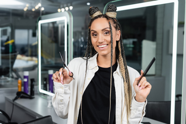 professional headshots, hairstylist with braids holding hairdressing scissors and comb in beauty salon, beauty worker, salon job, beauty salon work, hair trends, haircut, looking at camera - Φωτογραφία, εικόνα