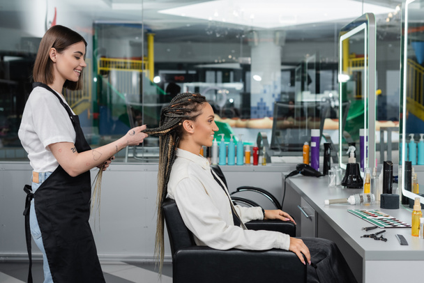 side view, client satisfaction, hairdresser styling hair of female customer, looking at mirror, happy woman with braids, hairstyle, braided hair, beauty salon, hairstyling products  - Photo, Image