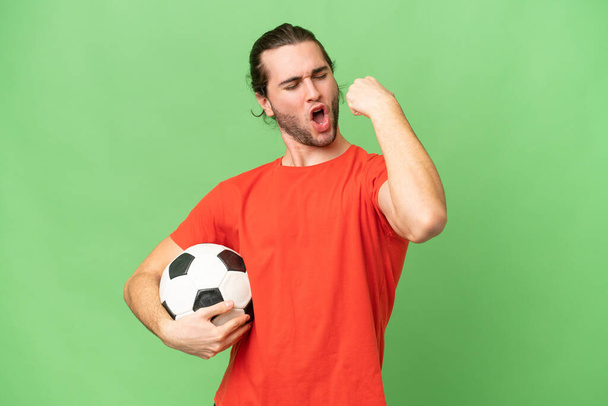 Молодой красивый мужчина, изолированный на зеленом фоне хромы с футбольным мячом, празднующим победу - Фото, изображение