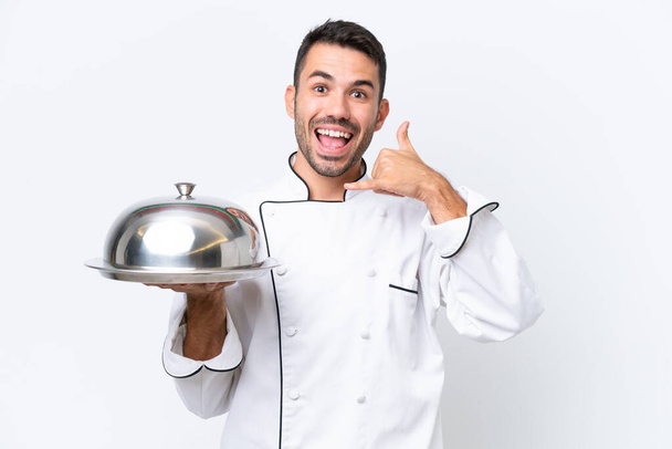 Young chef with tray isolated on white background making phone gesture. Call me back sign - Photo, Image