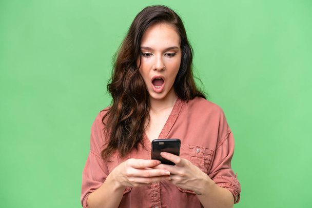 Young caucasian woman over isolated background looking at the camera while using the mobile with surprised expression - Photo, Image